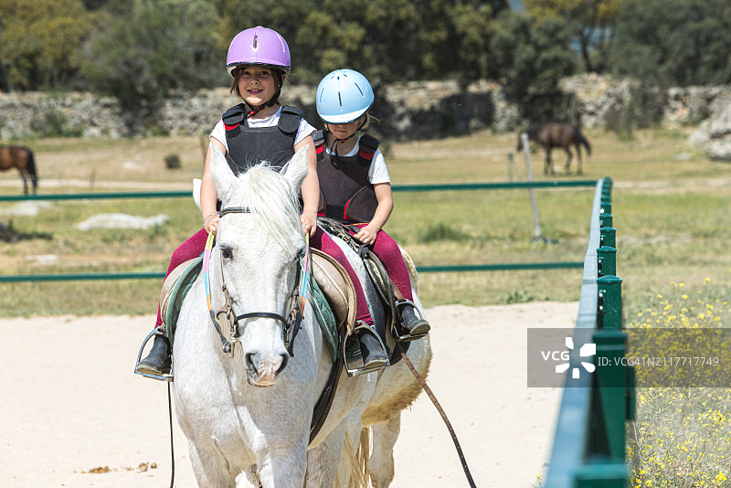 在骑术竞技场户外骑马的两个女孩的正面图：爱好、运动、马术、放松、健康、乡村、学习和自然图片素材