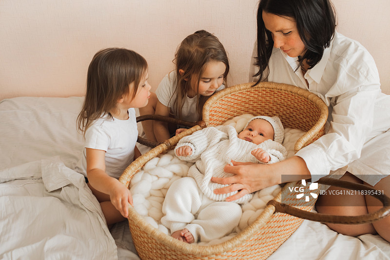 母亲和两个女儿在家中柳条婴儿床旁玩耍，照看新生弟弟图片素材