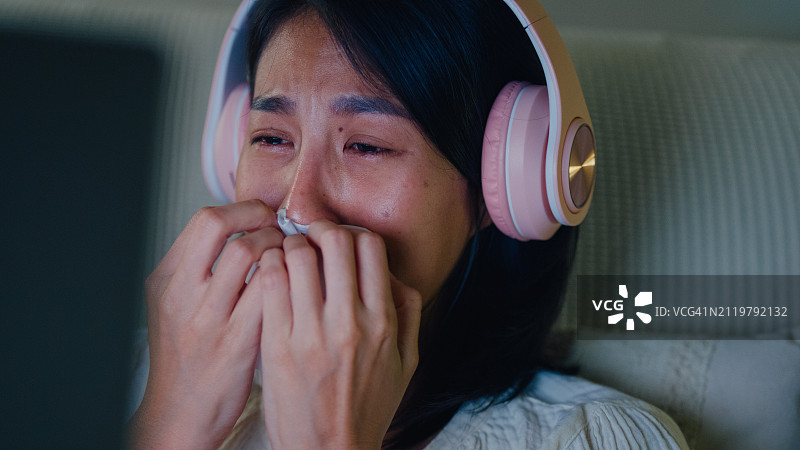 年轻亚洲女孩在床上戴着耳机哭泣，观看流媒体在线视频点播电影特写。失眠、网络病、睡眠障碍图片素材