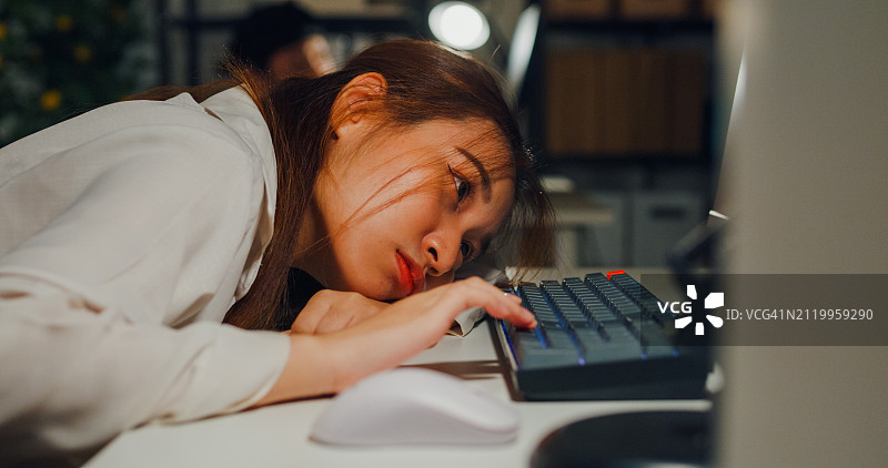 亚洲女性在办公室夜晚使用笔记本电脑时显得疲惫和有压力，突出了过度工作和员工倦怠的问题。ESG可持续商业办公室。图片素材