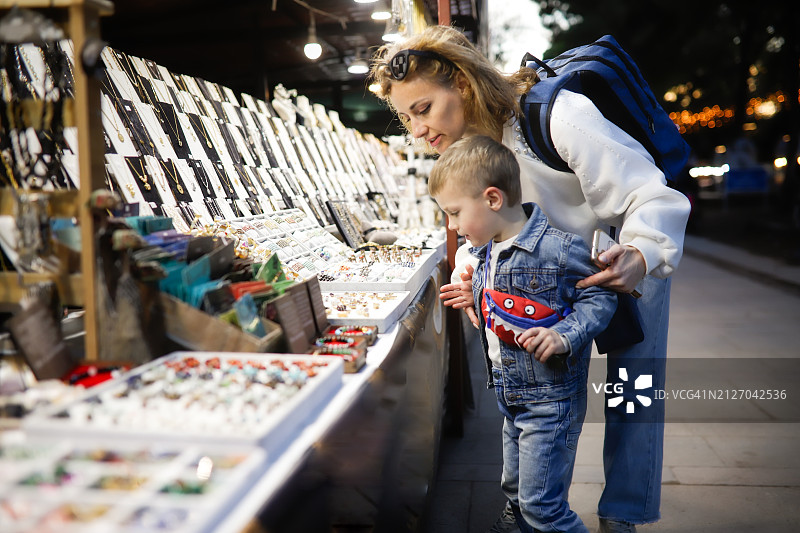家庭纪念品选购：中年妈妈和5岁儿子在夜市上浏览特色珠宝和纪念品，与孩子们一起体验城市旅行图片素材