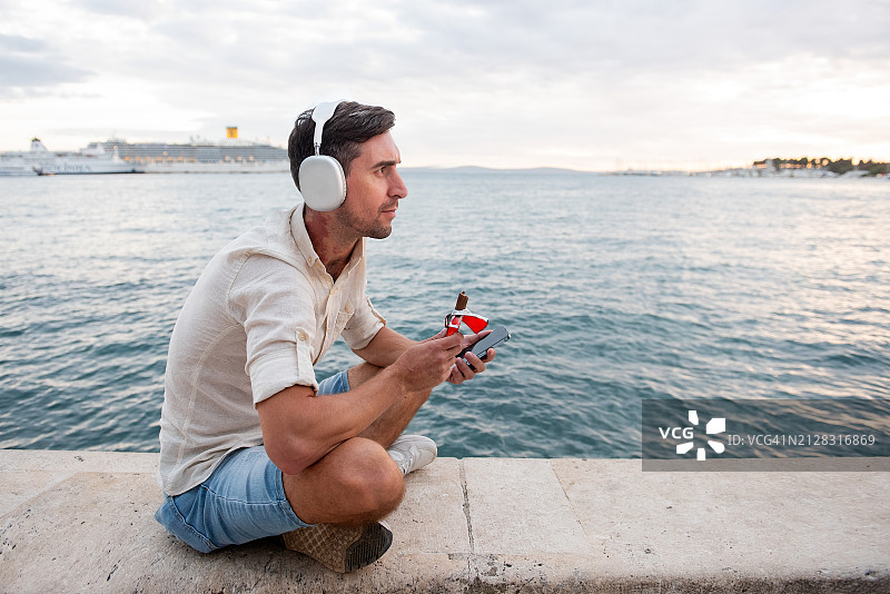 戴着大银色耳机，脖子上带着红色胎记的年轻男人在海滨长廊吃巧克力图片素材