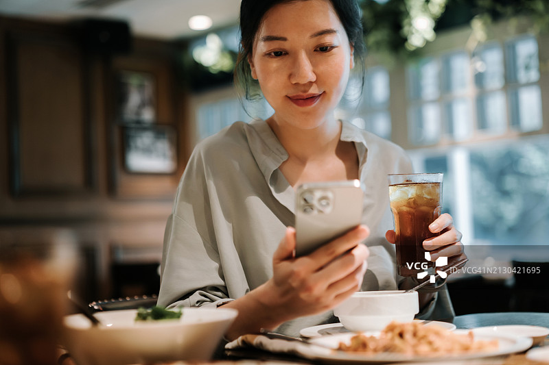 年轻亚洲女性在餐厅用餐时使用手机，享受午休时光。人物、美食、生活方式与科技图片素材