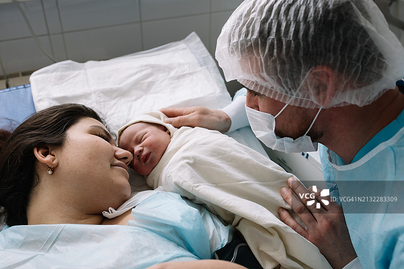 分娩后，母亲休息，看着新生婴儿，父亲坐在他们旁边分享特别的时刻图片素材