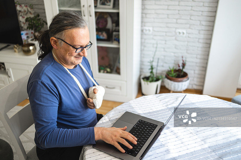 在家中使用笔记本电脑，手臂骨折打着石膏的中年白人男子图片素材