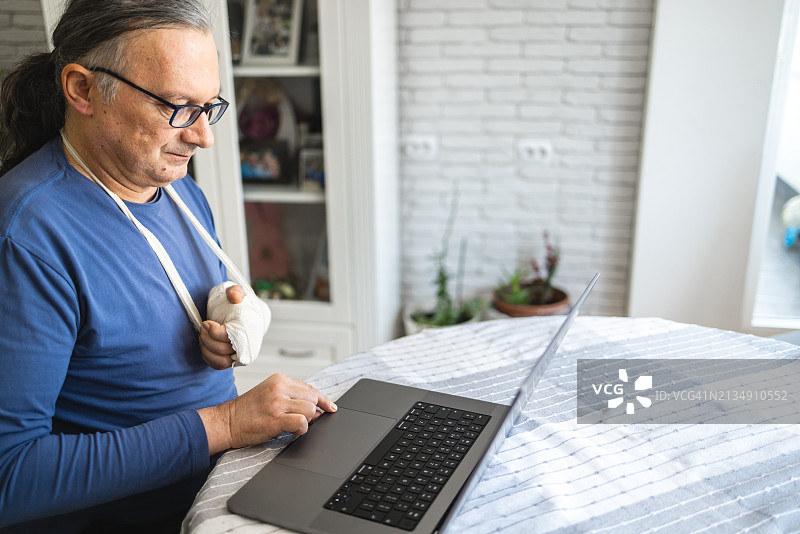 在家中使用笔记本电脑，手臂骨折打着石膏的中年白人男子图片素材