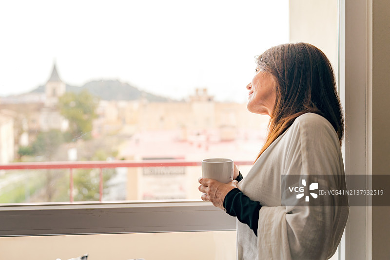 中年妇女在窗边品尝新鲜煮好的咖啡，欣赏美景图片素材