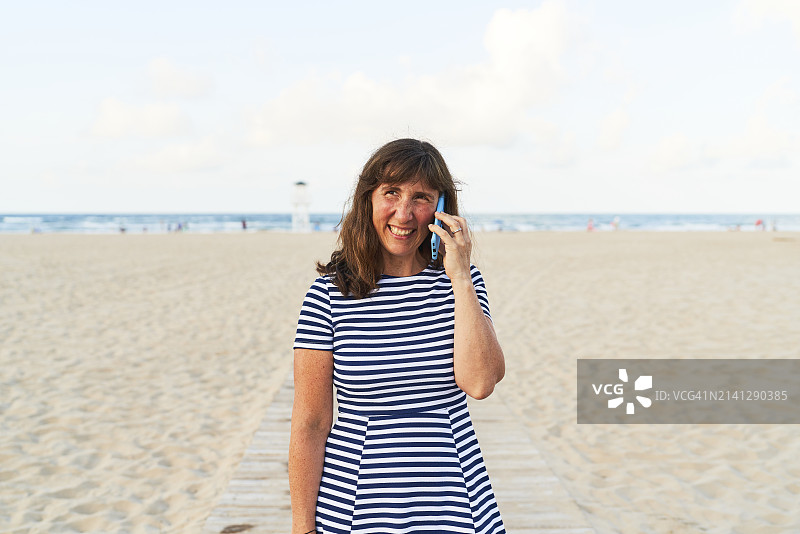 女子喜悦地通电话，身穿条纹连衣裙，背景是模糊的沙滩图片素材