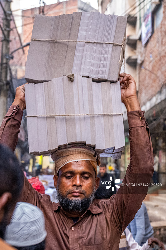 在达卡，一位身着传统服装的男人扛着一叠厚厚的纸板穿过拥挤的街道，体现了这座城市的韧性和勤劳精神。图片素材