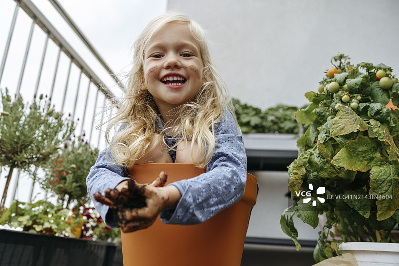 快乐的女孩双手脏兮兮地坐在阳台植物旁的花盆里图片素材