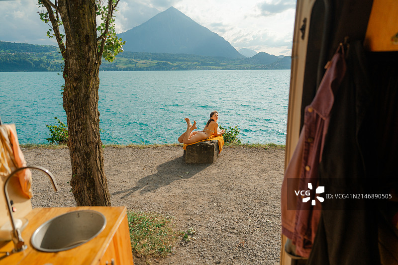女人在岩石上晒日光浴，在山间湖泊附近读书，从露营车可见图片素材