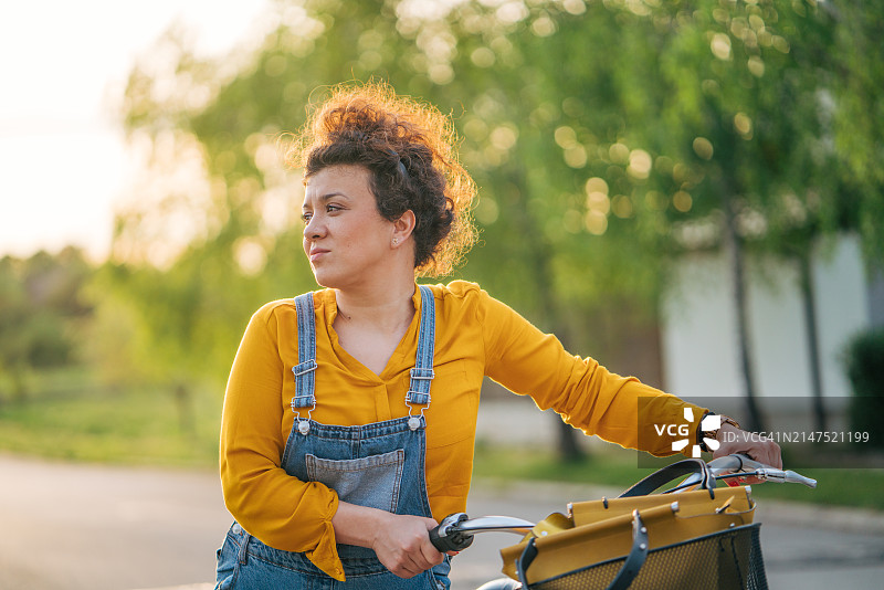 在阳光明媚的公园里推着自行车的卷发微笑女性图片素材