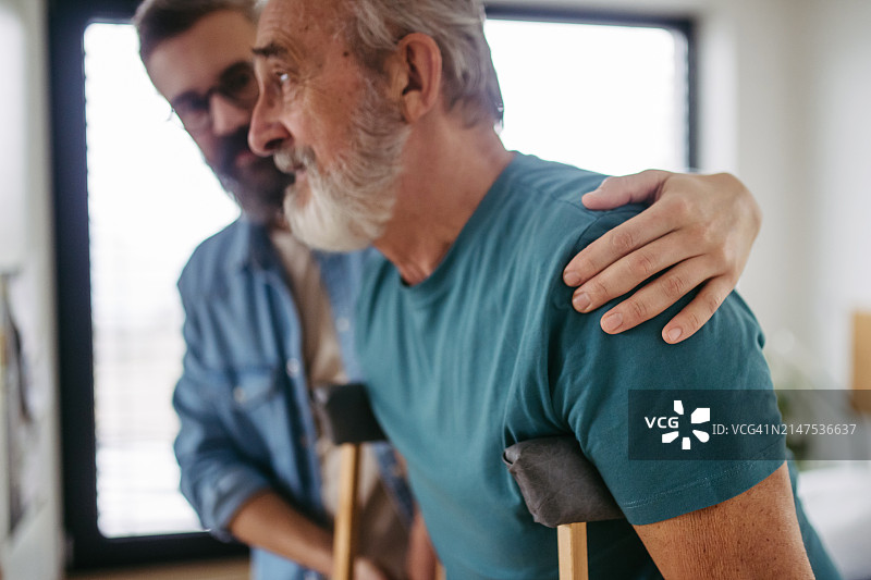 男子帮助拄拐杖的老年父亲，术后康复，儿子帮助父亲走路图片素材