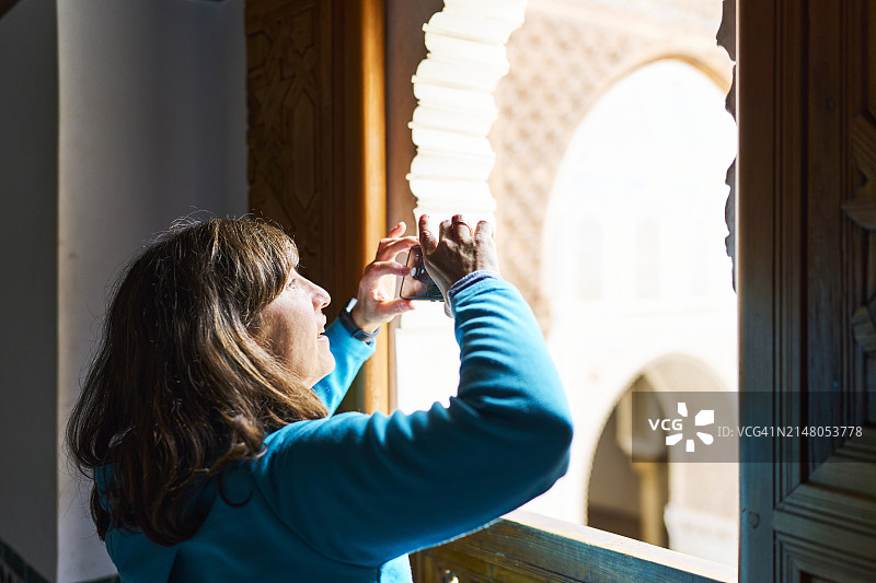 身穿休闲装站在马拉喀什宫殿内的女游客透过窗户拍照的侧面画像图片素材