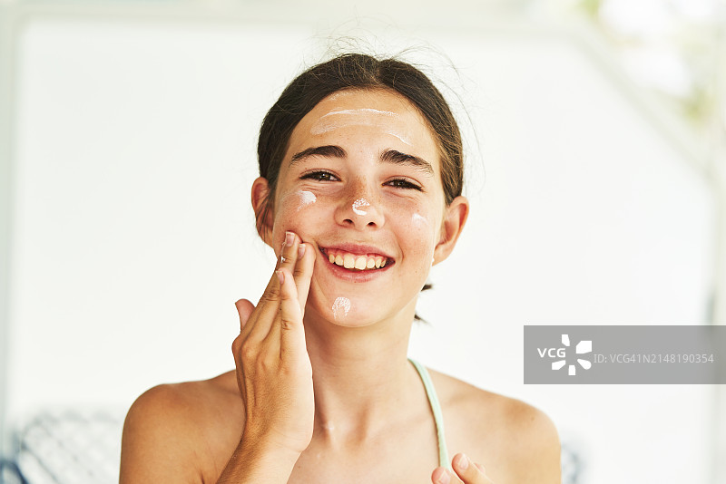 一位美丽的年轻女人在脸颊上涂抹面霜，面带微笑地看着镜头，正面视角图片素材