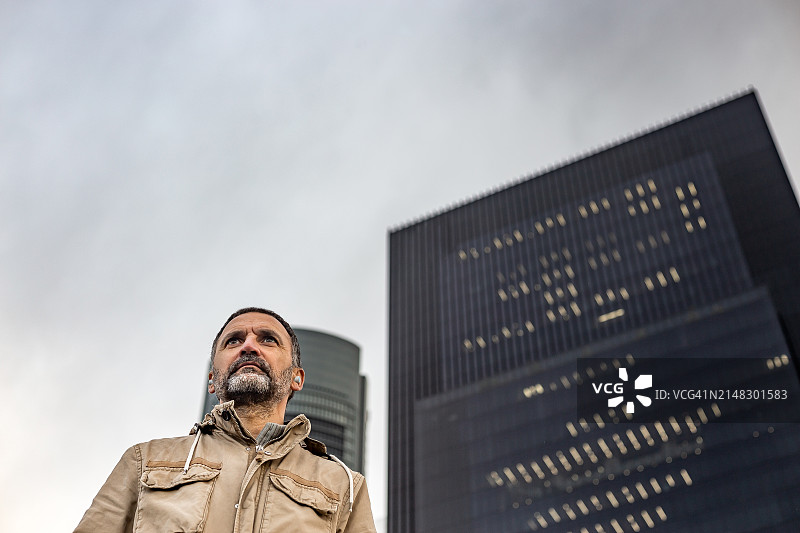 金融区摩天大楼背景下，戴着入耳式耳机沉思的成熟男人眺望远方的低角度仰视图片素材