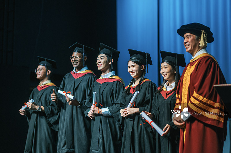 毕业典礼上自豪的亚洲中国毕业生从院长手中接过文凭，在礼堂舞台上看着镜头微笑图片素材