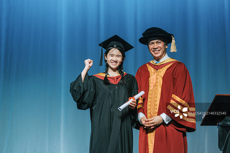 毕业典礼：兴奋的亚洲女大学生在礼堂舞台上收到毕业证书后，站在院长旁边举起手臂图片素材