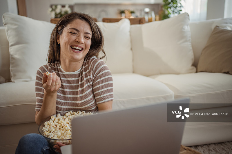 年轻女人坐在家里的地板上靠在沙发上看喜剧电影图片素材