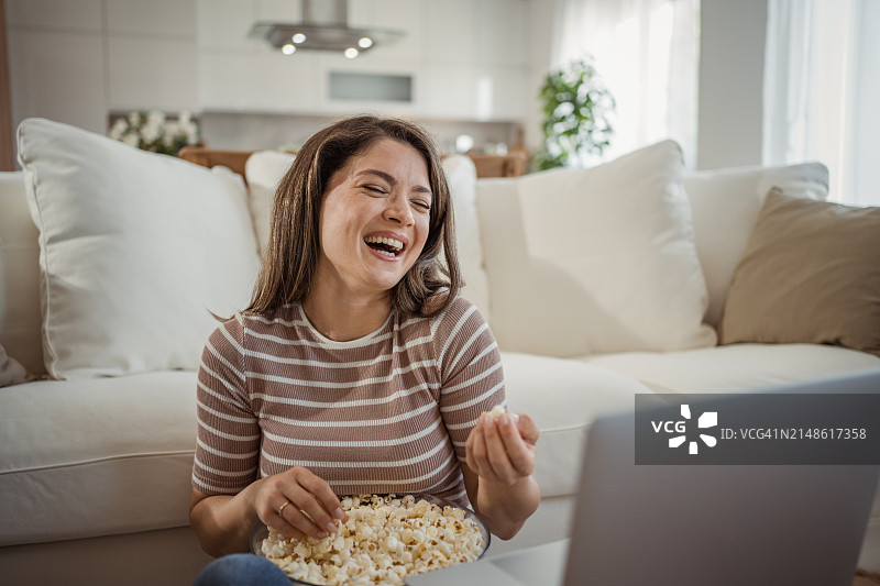 年轻女人坐在家里的地板上靠在沙发上看喜剧电影图片素材