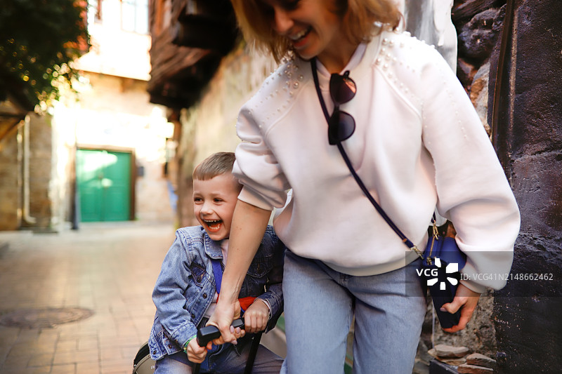 快乐旅程：中年母亲在老城街道上趣味地骑着坐在行李箱上的5岁儿子图片素材