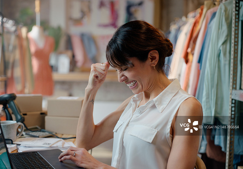 时装设计师在笔记本电脑上获得成功，庆祝电子商务，并对销售额、利润或贷款批准感到兴奋。小企业主或有服装订单、库存或客户评价的女性图片素材