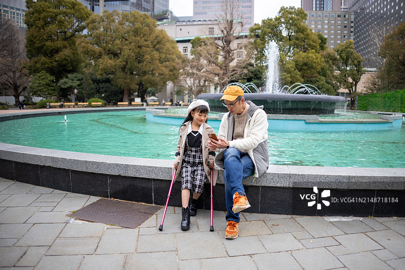 在公园喷泉前与父亲同坐的拄拐杖小女孩图片素材