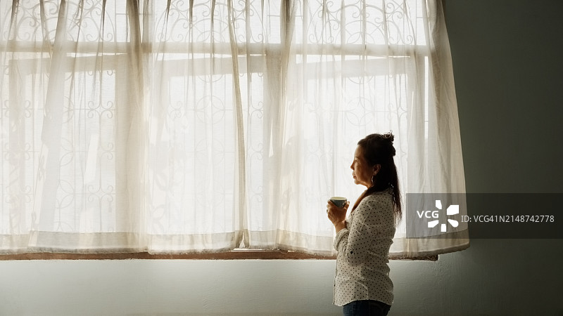 手持咖啡杯站在落地窗前的亚洲老年妇女图片素材