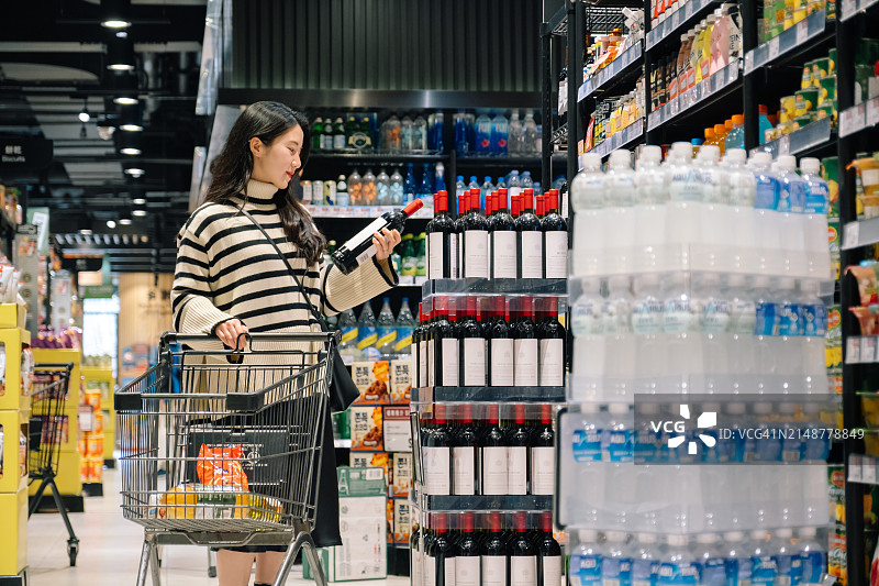 年轻的亚洲女人在超市货架上挑选一瓶红酒。图片素材