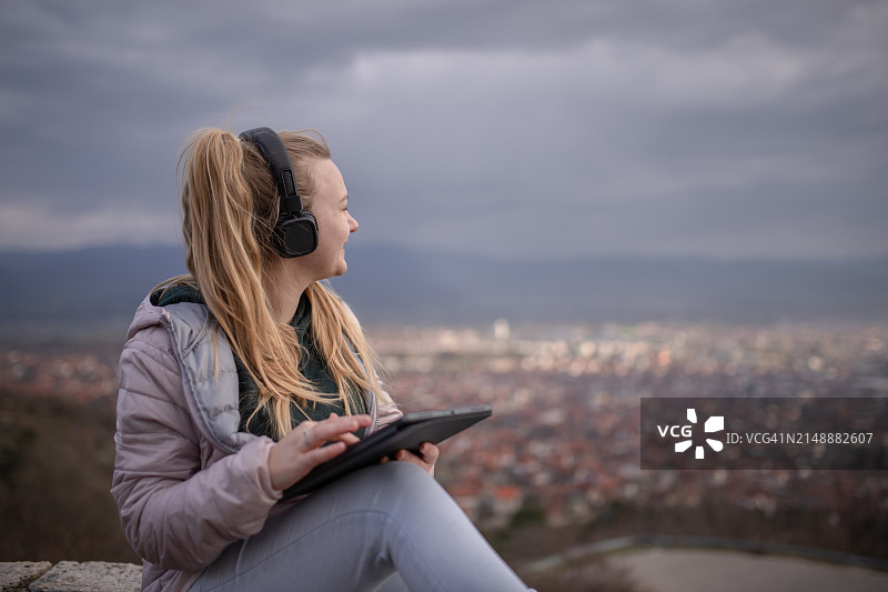 戴着耳机坐在城市上空，使用平板电脑的金发年轻女子图片素材