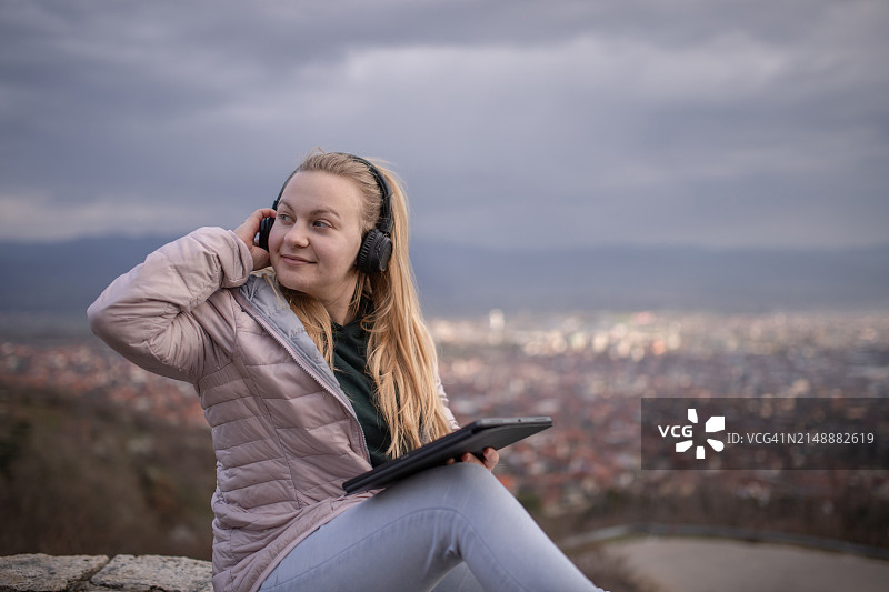 年轻金发女子戴着耳机坐在城市上空，使用平板电脑听音乐图片素材