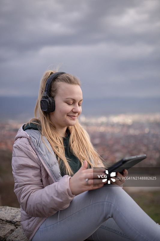 戴着耳机坐在城市上空，使用平板电脑的金发年轻女子图片素材