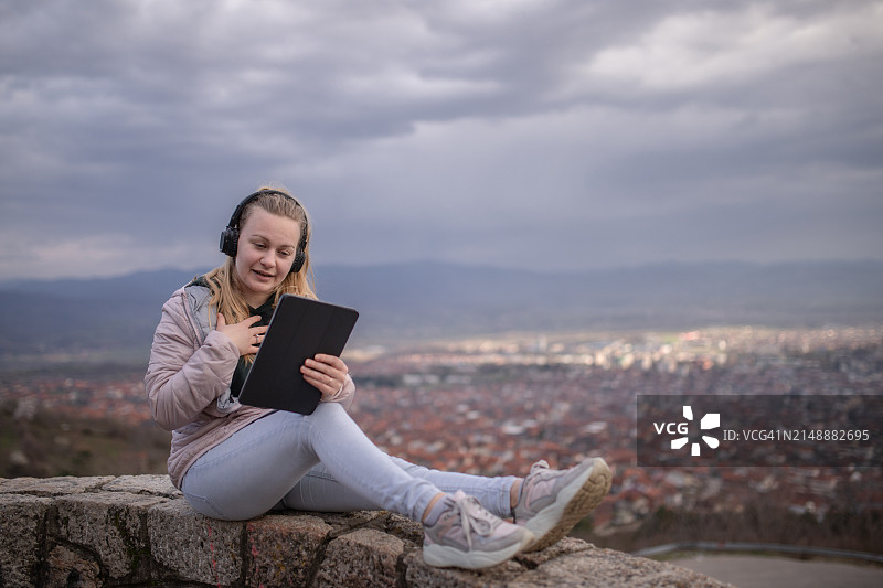 戴着耳机坐在城市上空，使用平板电脑的金发年轻女子图片素材
