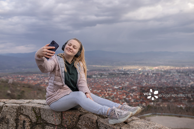 年轻金发女子戴耳机在城市上空用智能手机自拍图片素材