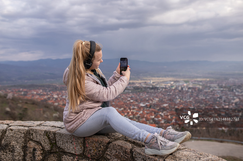 戴着耳机坐在城市上空，用智能手机拍摄城市美景的金发年轻女子图片素材