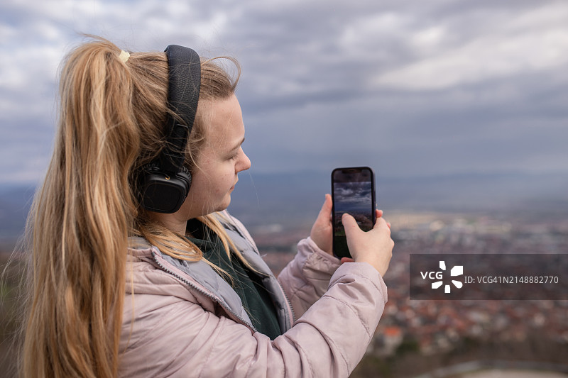 戴着耳机坐在城市上空，用智能手机拍摄城市美景的金发年轻女子图片素材