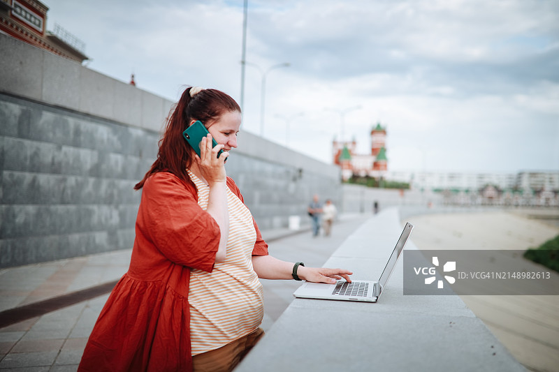 城市工作：中年妇女在街头水泥堤岸上用笔记本电脑和手机处理多项任务图片素材