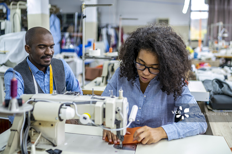 非裔美国青年同事在服装制造工作室讨论，女性在现代缝纫机上缝纫图片素材
