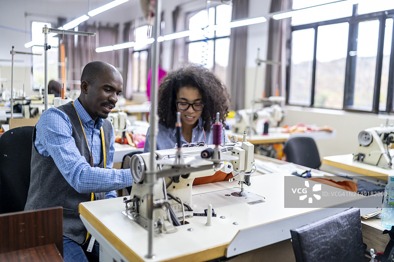 非裔美国青年同事在服装制造工作室讨论，女性在现代缝纫机上缝纫图片素材
