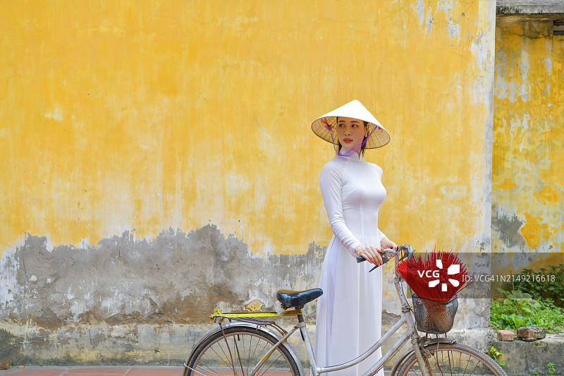 身穿传统奥黛，站在越南河内黄色墙前的越南女子和她的自行车图片素材
