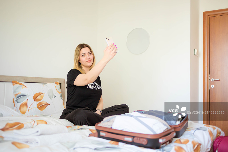 快乐的女人坐在床上，一边收拾行李箱一边使用手机图片素材