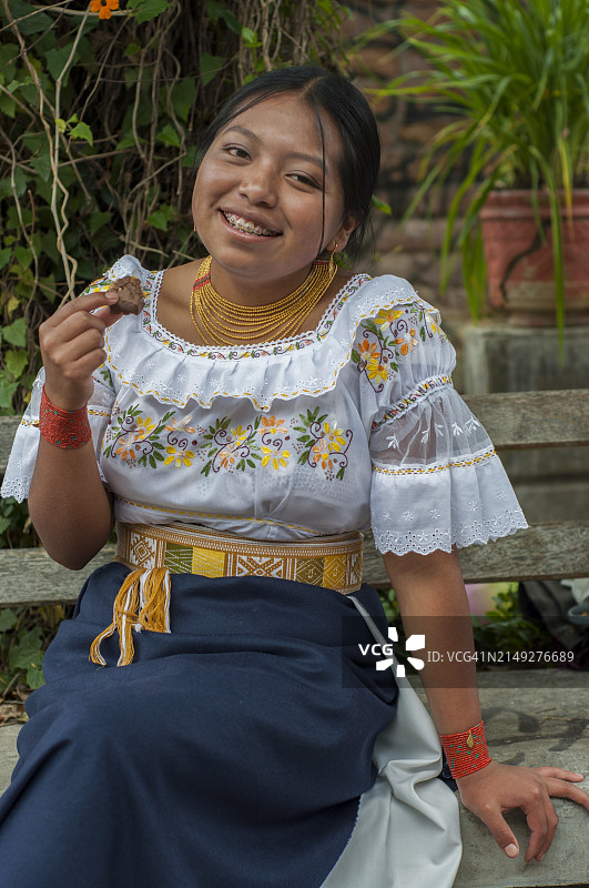 厄瓜多尔奥塔瓦洛一位身穿手工刺绣衬衫和裙子的快乐年轻女子坐在公园里品尝巧克力糖果。图片素材