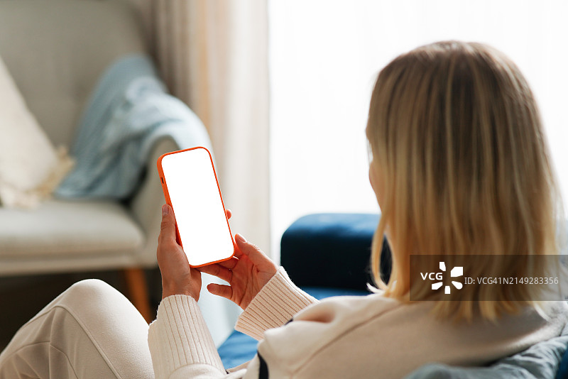 一位穿着毛衣的金发女人坐在家里的蓝色沙发上，用白色屏幕的手机聊天图片素材