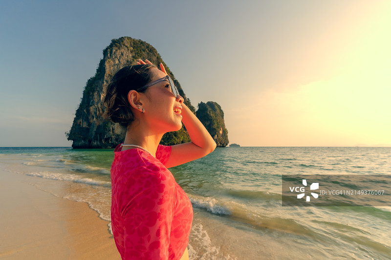 穿着红色比基尼和太阳镜的女人在金色日落时分走在沙滩上，背景是山脉图片素材