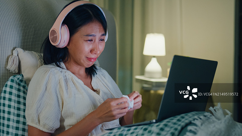 年轻的亚洲女孩在床上戴着耳机哭着观看在线视频，沉迷于媒体，失眠图片素材