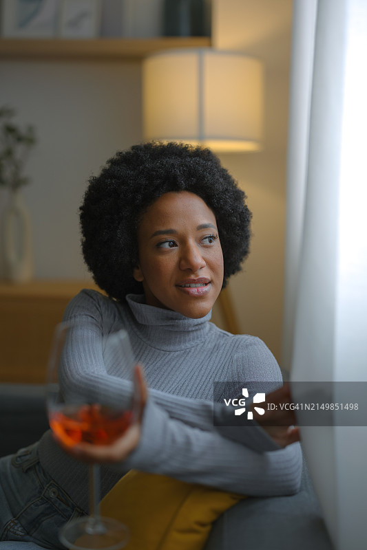 年轻拉丁裔女人在现代公寓舒适的沙发上放松，一边品尝红酒，一边眺望窗外图片素材