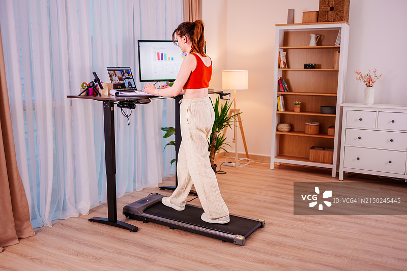 在符合人体工程学的站立式办公桌上工作的年轻女商人，在跑步机上行走，在线会议时查看数字交易所上的比特币或股市价格图表图片素材