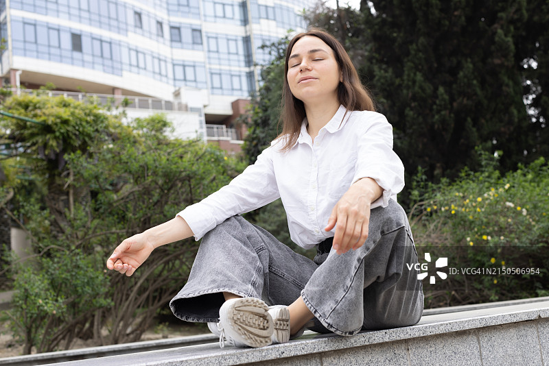 一位美丽的年轻女子身穿牛仔裤和白衬衫，坐在街边，以高楼大厦为背景图片素材