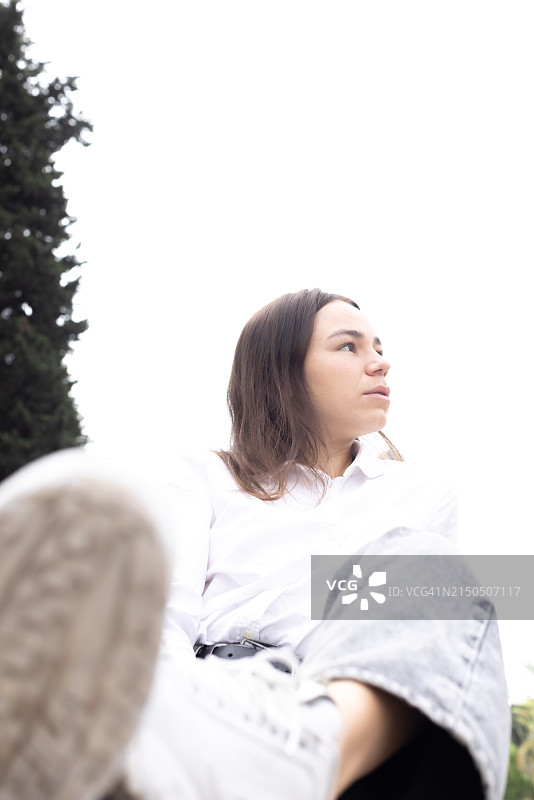 一位美丽的年轻女子身穿牛仔裤和白衬衫，坐在街边，以高楼大厦为背景图片素材