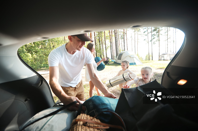 家庭露营：快乐的孩子们在河边的松树林里帮助父母打开汽车图片素材